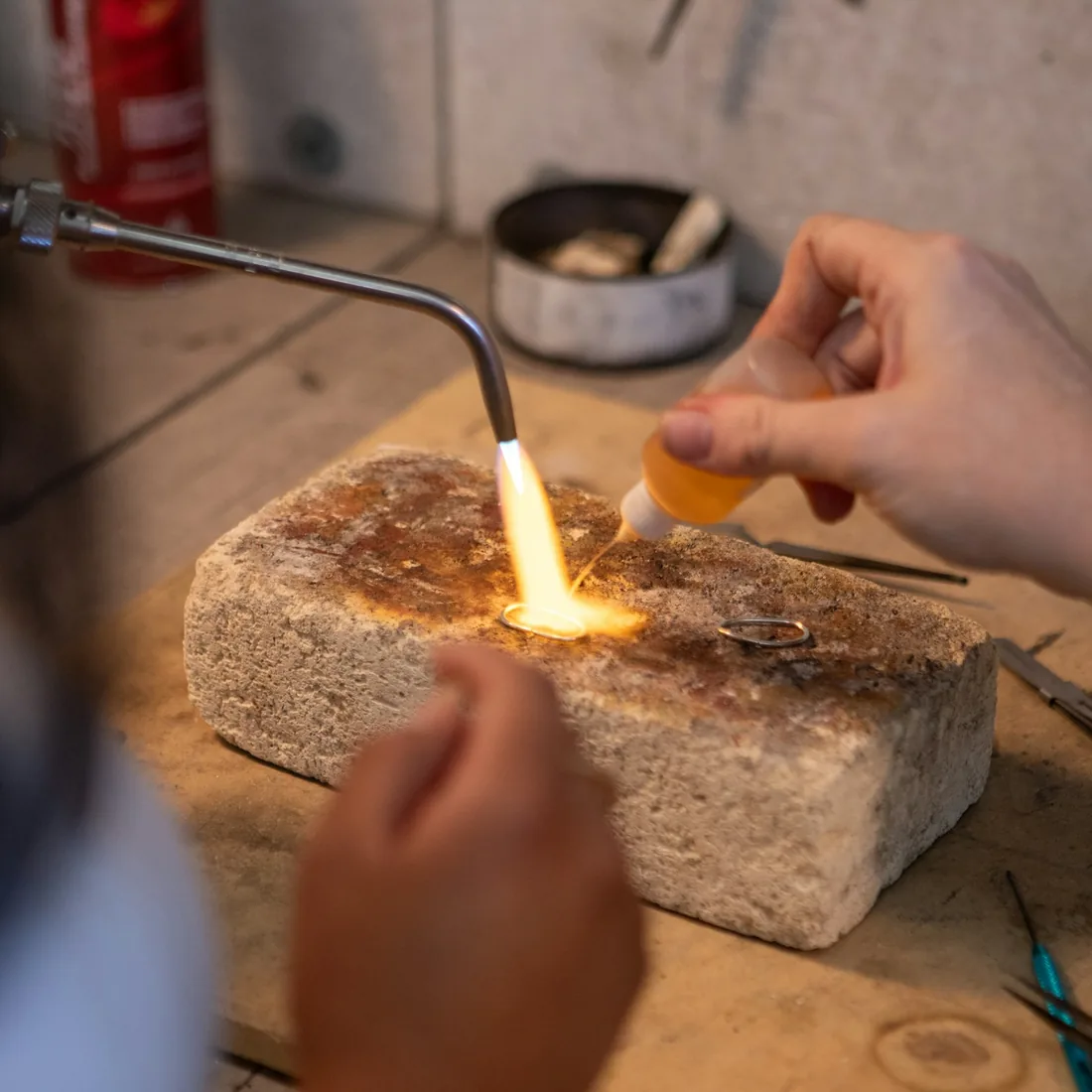 Soplete encendido sobre un ladrillo refractario en el banco de un taller de metal, con pinzas y herramientas alrededor
